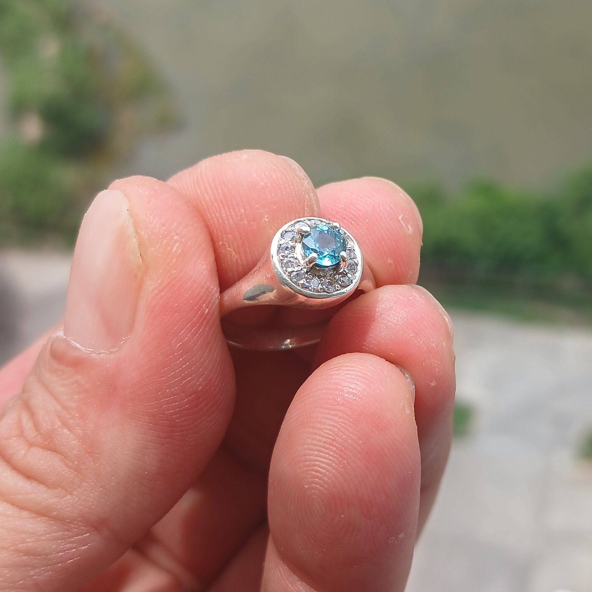 A stunning Blue Zircon Ring featuring a natural blue Zircon stone surrounded by 13 white CZ stones, set in polished sterling silver.