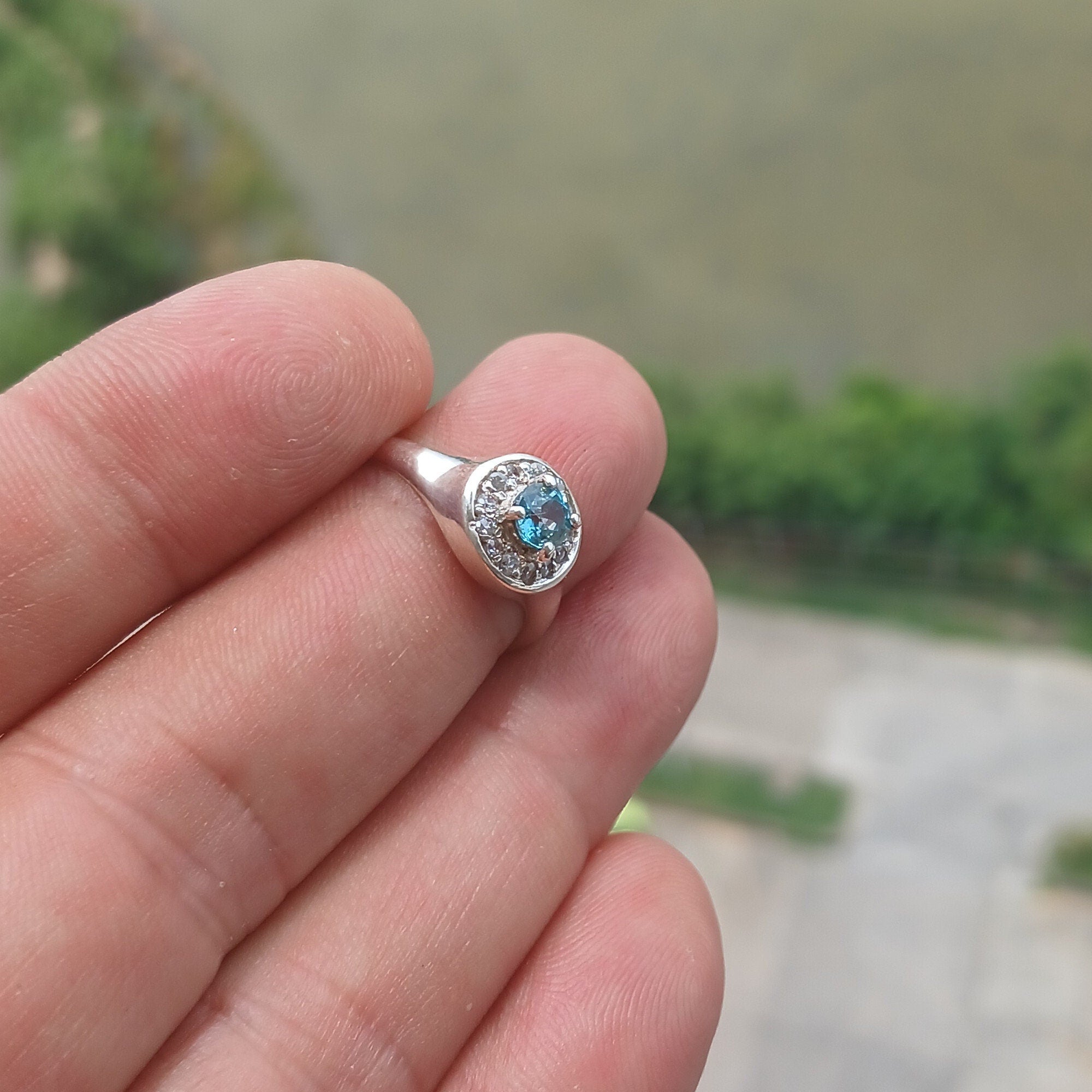 A stunning Blue Zircon Ring featuring a natural blue Zircon stone surrounded by 13 white CZ stones, set in polished sterling silver.