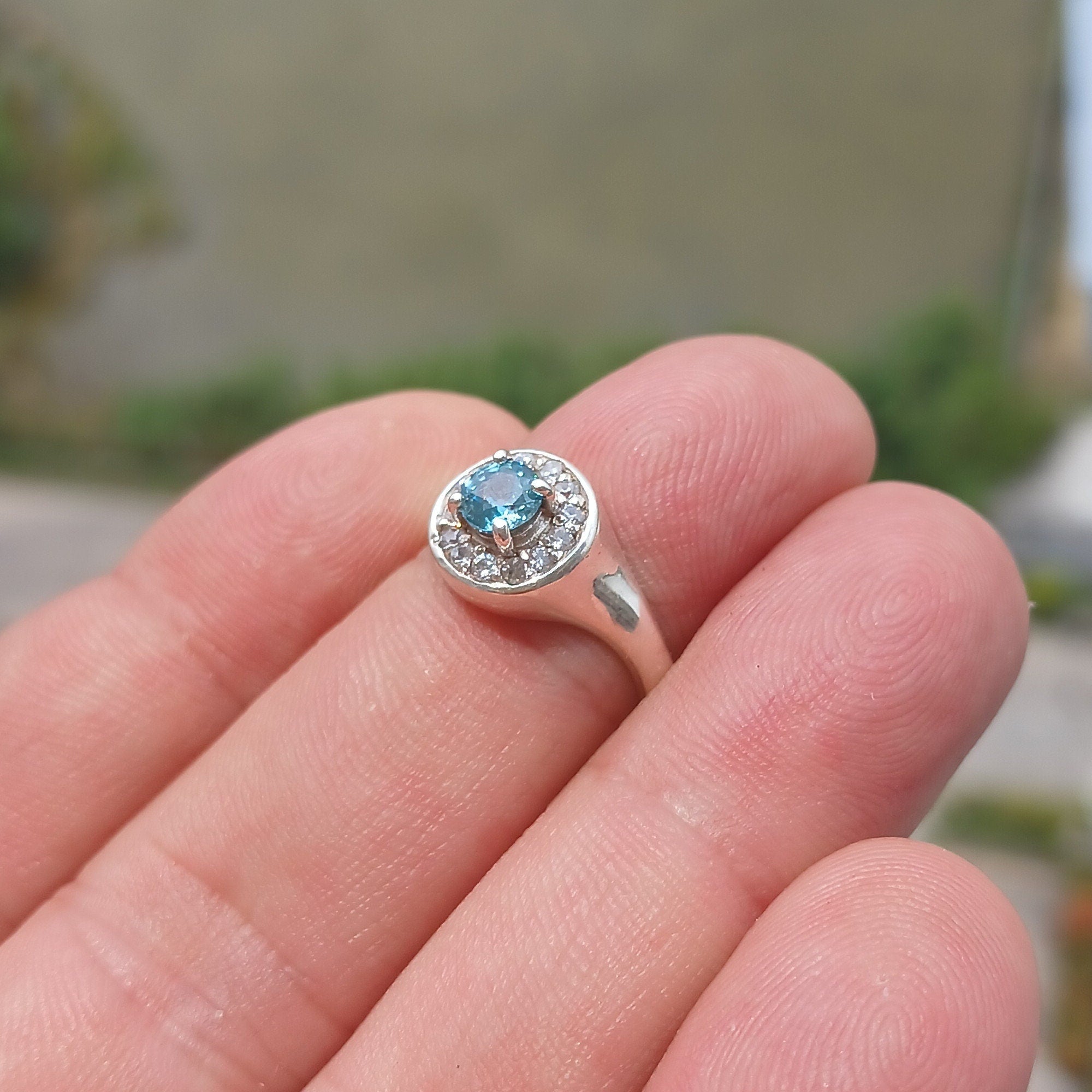 A stunning Blue Zircon Ring featuring a natural blue Zircon stone surrounded by 13 white CZ stones, set in polished sterling silver.