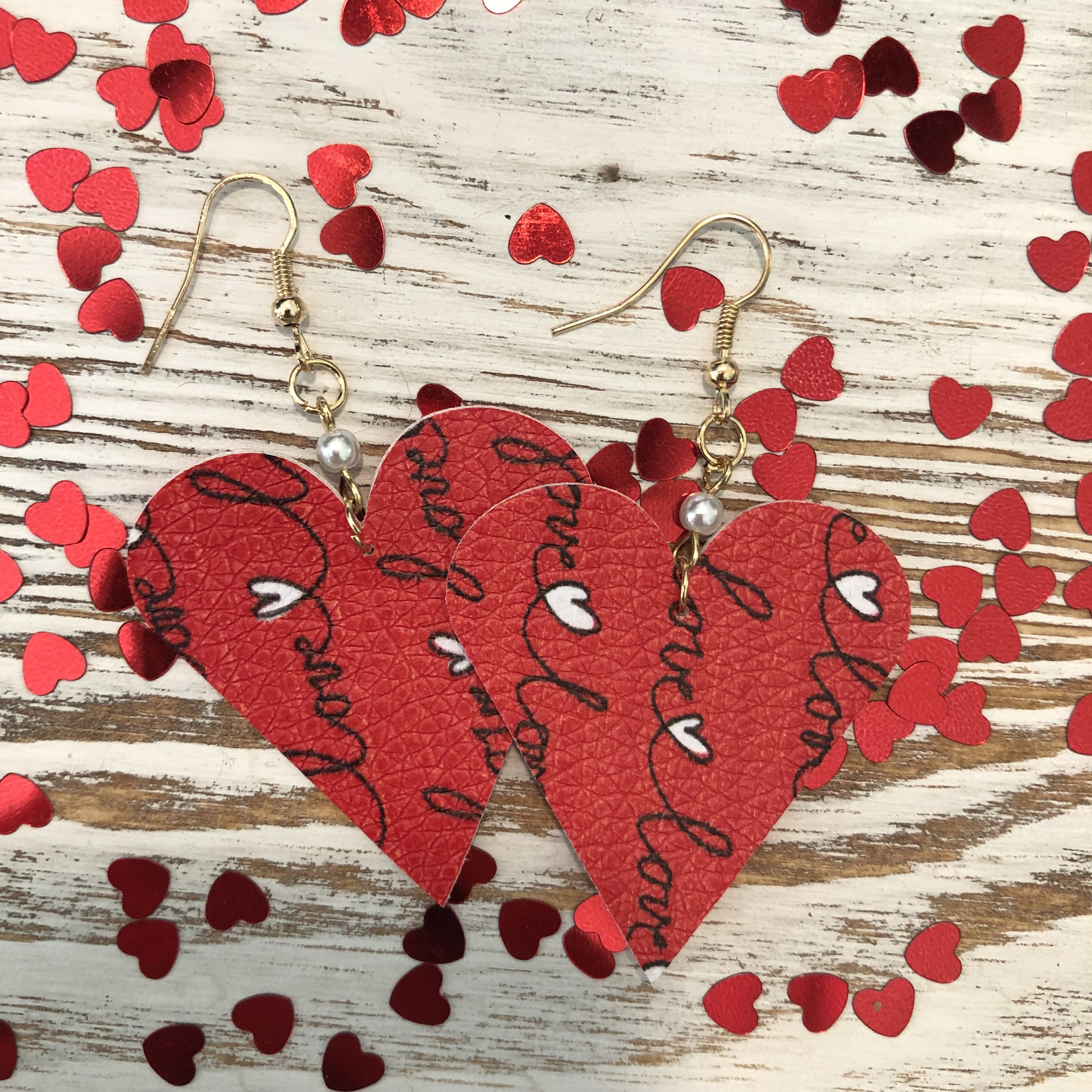 A pair of lightweight red heart-shaped leather earrings with pearl accents, featuring a fish hook design and double-sided style.