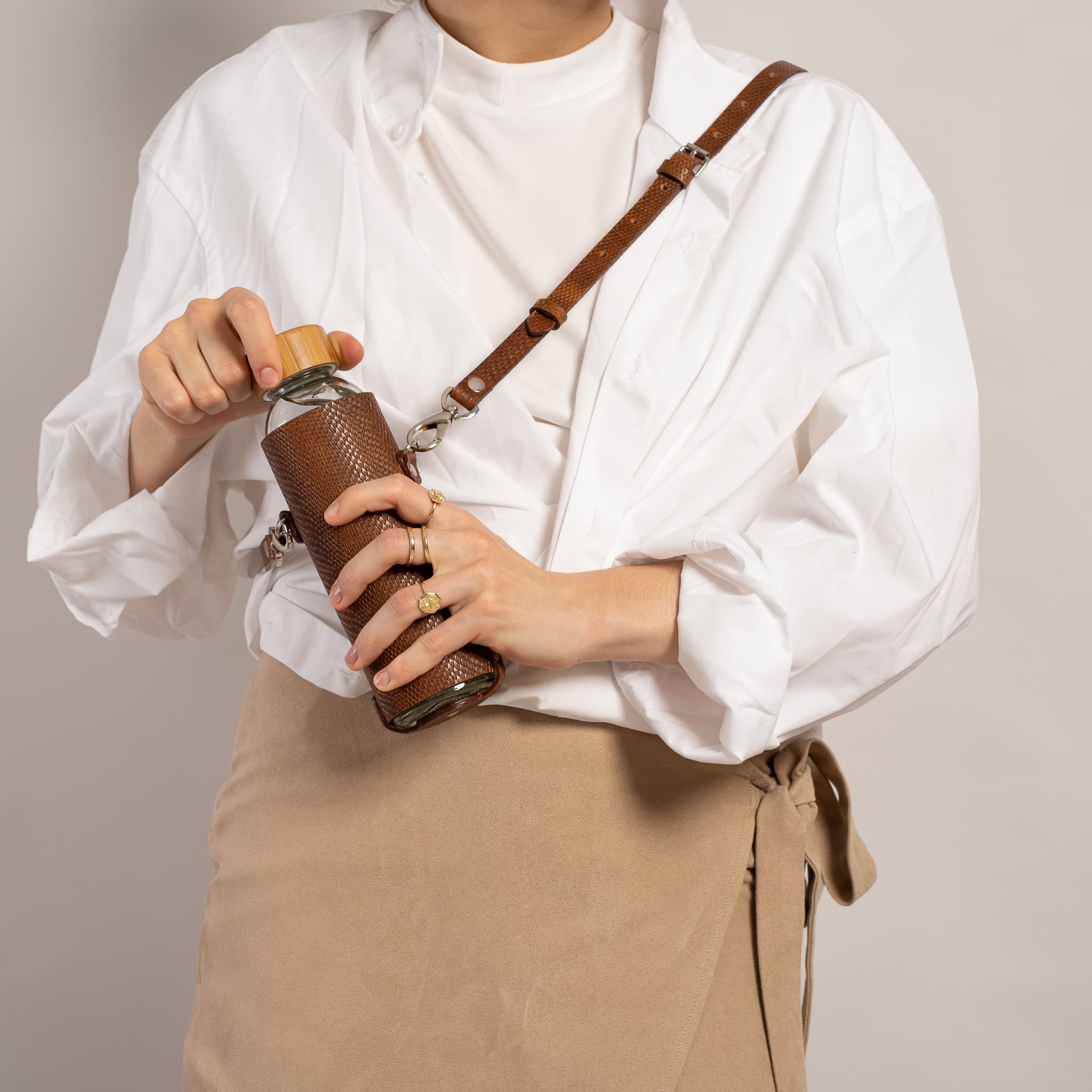 Leather water bottle holder featuring a glass bottle, bamboo lid, and adjustable strap, designed for stylish hydration on the go.