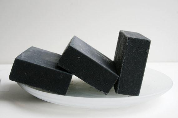 A bar of Activated Charcoal Soap showcasing its dark color and natural texture, surrounded by essential oils and natural ingredients, emphasizing its handmade quality.