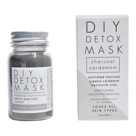 A jar of Charcoal Cardamom Detox Face Mask with a wooden spoon, showcasing its rich black color and texture, surrounded by cardamom pods and activated charcoal.