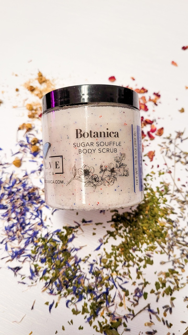 A jar of Botanica Sugar Souffle Body Polish with a botanical garden background, showcasing its creamy texture and luxurious packaging.