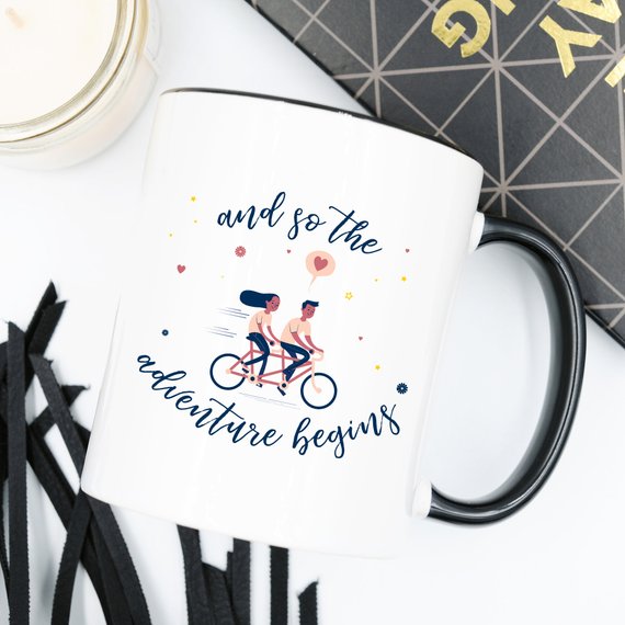 A ceramic coffee mug featuring the phrase 'And So The Adventure Begins' in a fun, colorful design, perfect for coffee lovers and adventurers.