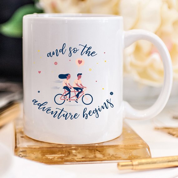 A ceramic coffee mug featuring the phrase 'And So The Adventure Begins' in a fun, colorful design, perfect for coffee lovers and adventurers.