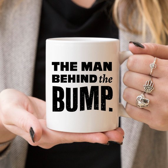 A humorous coffee mug featuring the phrase 'The Man Behind The Bump', crafted from high-grade ceramic, perfect for gifting.