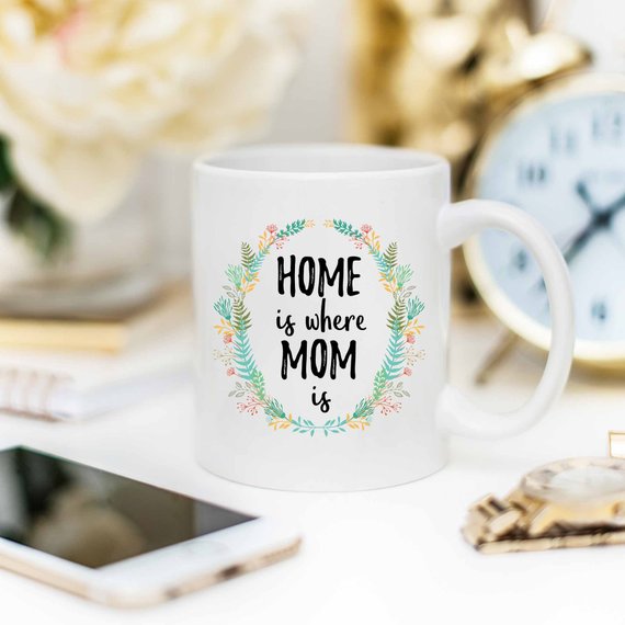 A white ceramic mug with the phrase 'Home Is Where Mom Is' printed in vibrant colors on both sides, showcasing a heartfelt design.