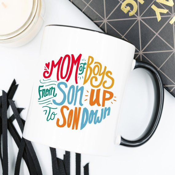 A humorous coffee mug designed for moms of boys, featuring the phrase 'Mom Of Boys, From Son Up To Son Down' in a playful font.
