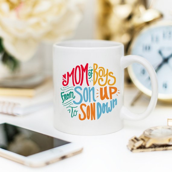 A humorous coffee mug designed for moms of boys, featuring the phrase 'Mom Of Boys, From Son Up To Son Down' in a playful font.