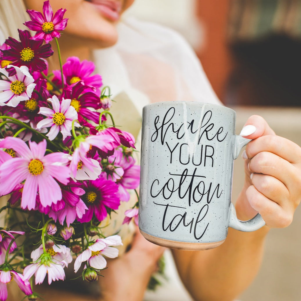 A pastel blue and pink ceramic coffee mug with 'Shake Your Cotton Tail' graphic, showcasing a fun Easter design.