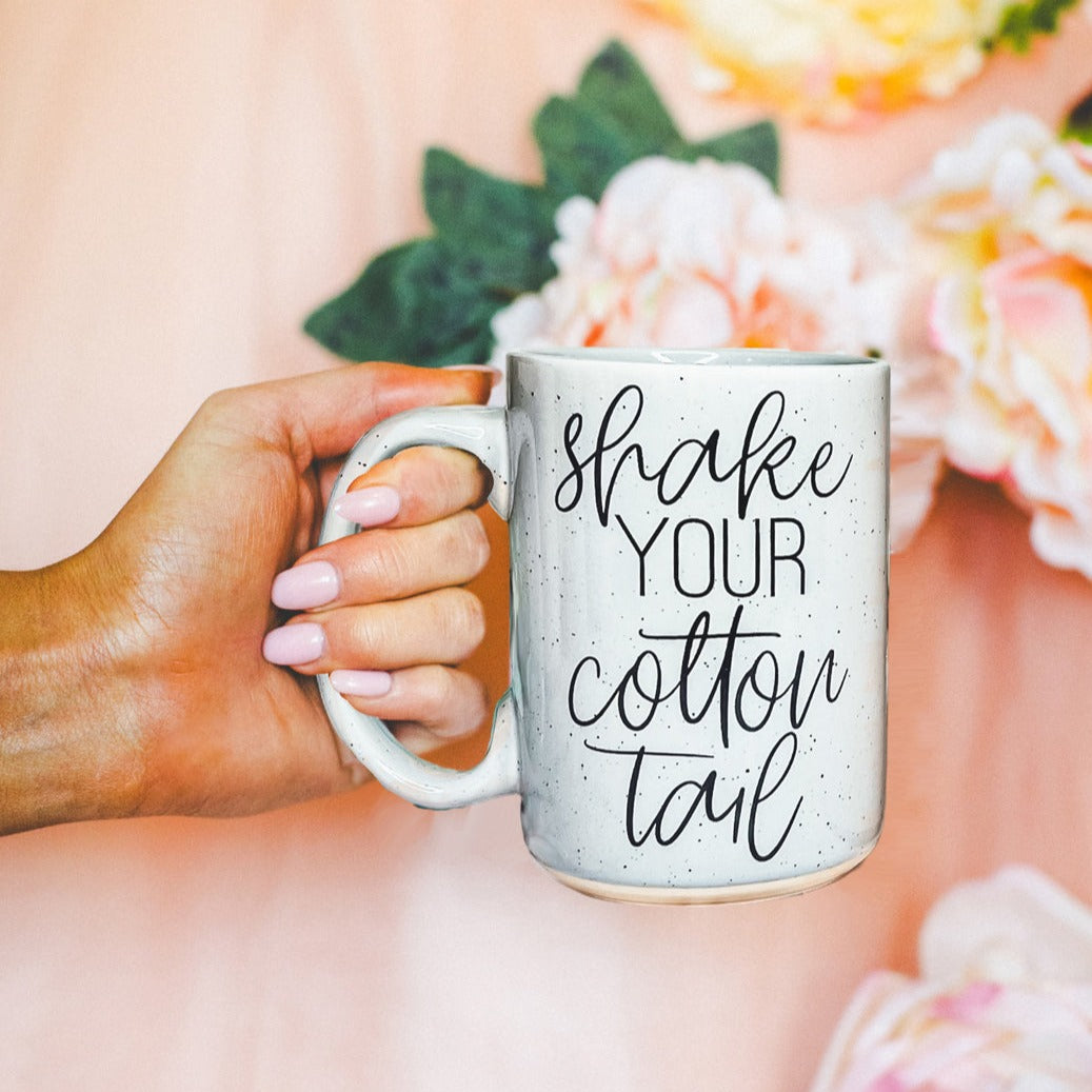 A pastel blue and pink ceramic coffee mug with 'Shake Your Cotton Tail' graphic, showcasing a fun Easter design.