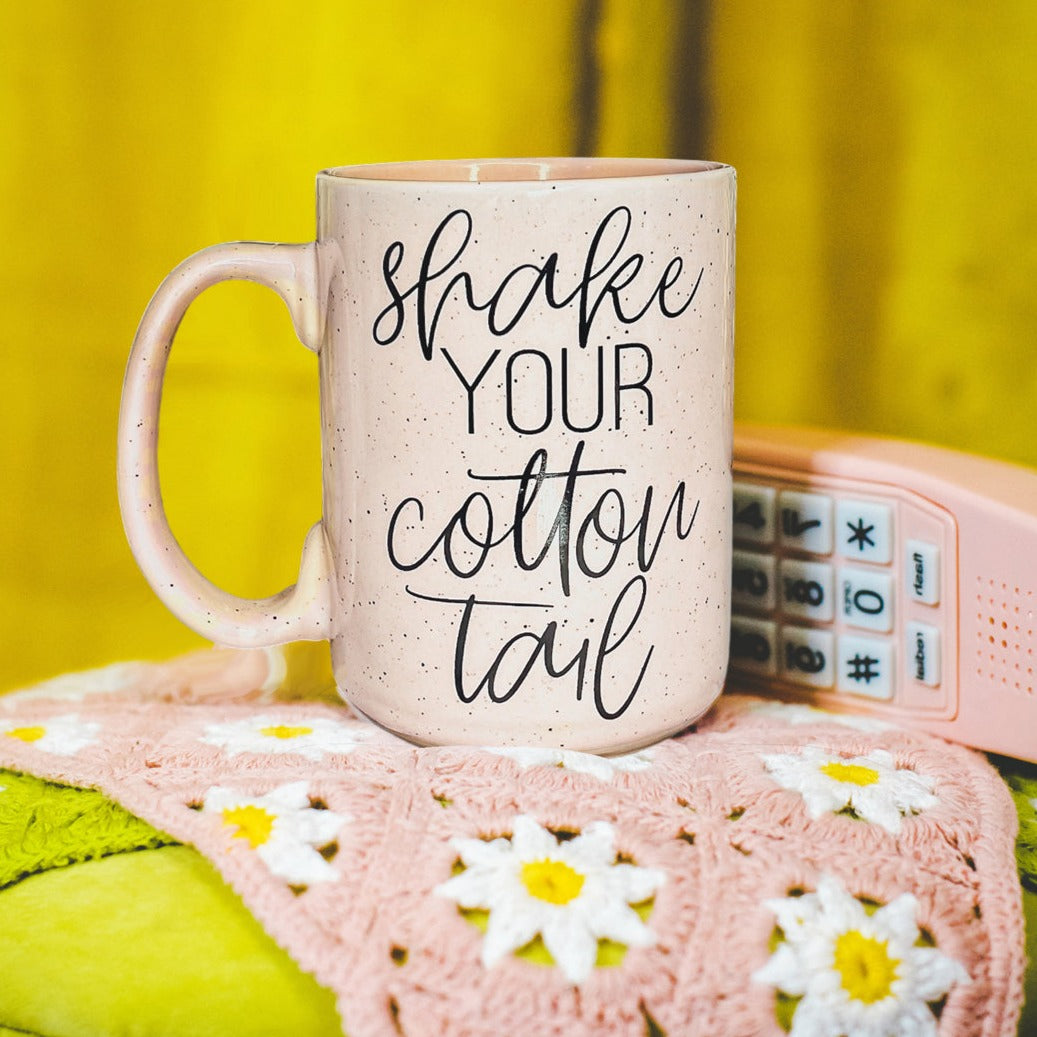 A pastel blue and pink ceramic coffee mug with 'Shake Your Cotton Tail' graphic, showcasing a fun Easter design.