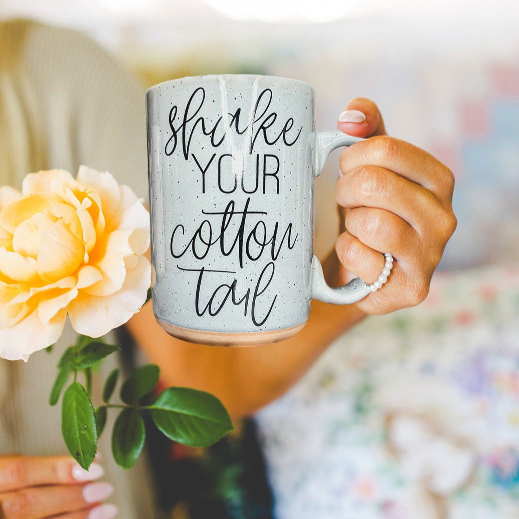 A pastel blue and pink ceramic coffee mug with 'Shake Your Cotton Tail' graphic, showcasing a fun Easter design.