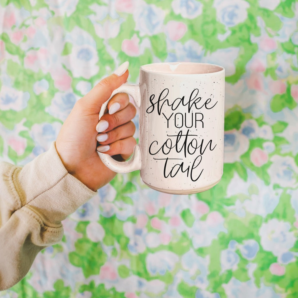 A pastel blue and pink ceramic coffee mug with 'Shake Your Cotton Tail' graphic, showcasing a fun Easter design.