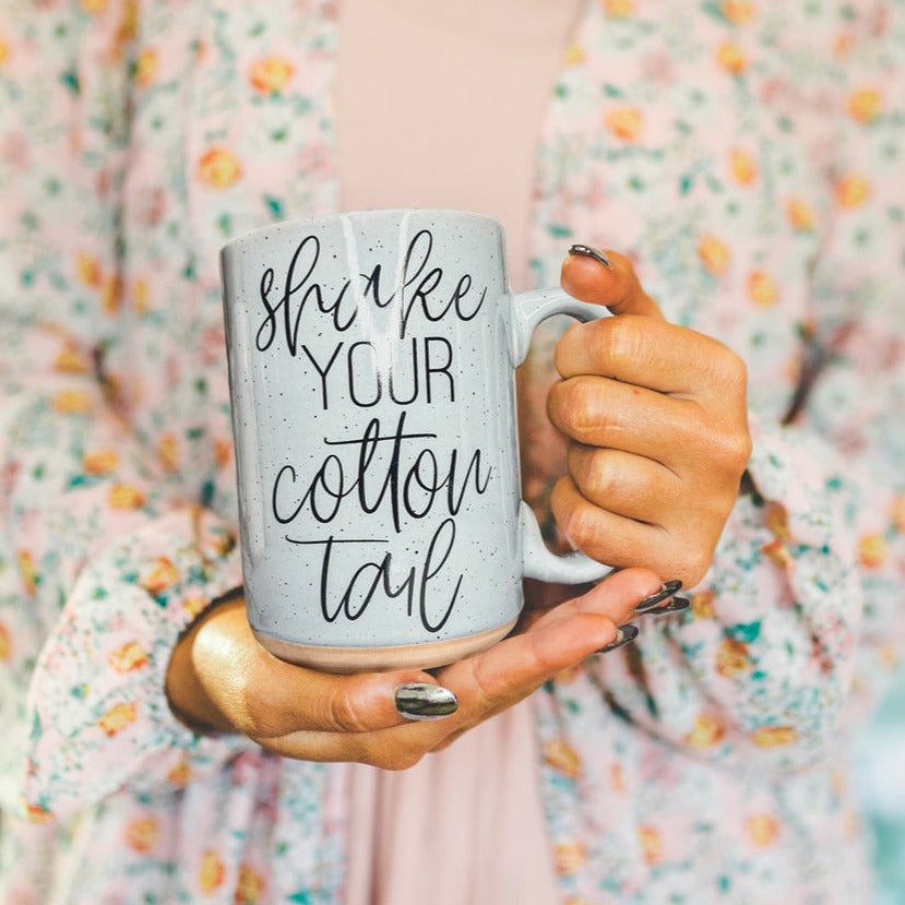 A pastel blue and pink ceramic coffee mug with 'Shake Your Cotton Tail' graphic, showcasing a fun Easter design.