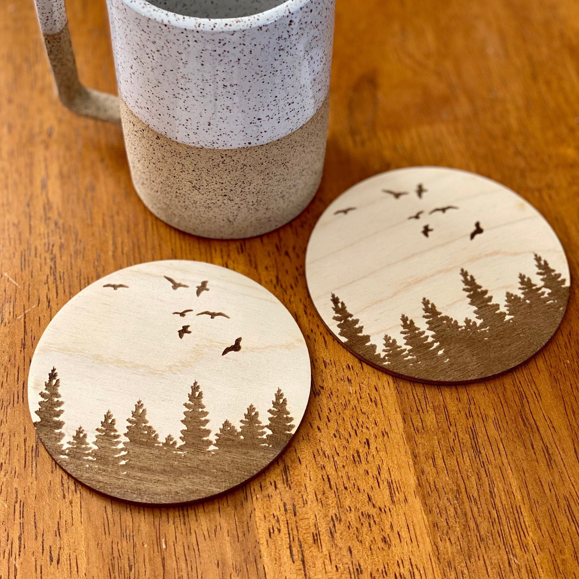 A set of four wooden coasters featuring engraved pine tree and bird designs, showcasing natural wood grain and a rustic aesthetic.