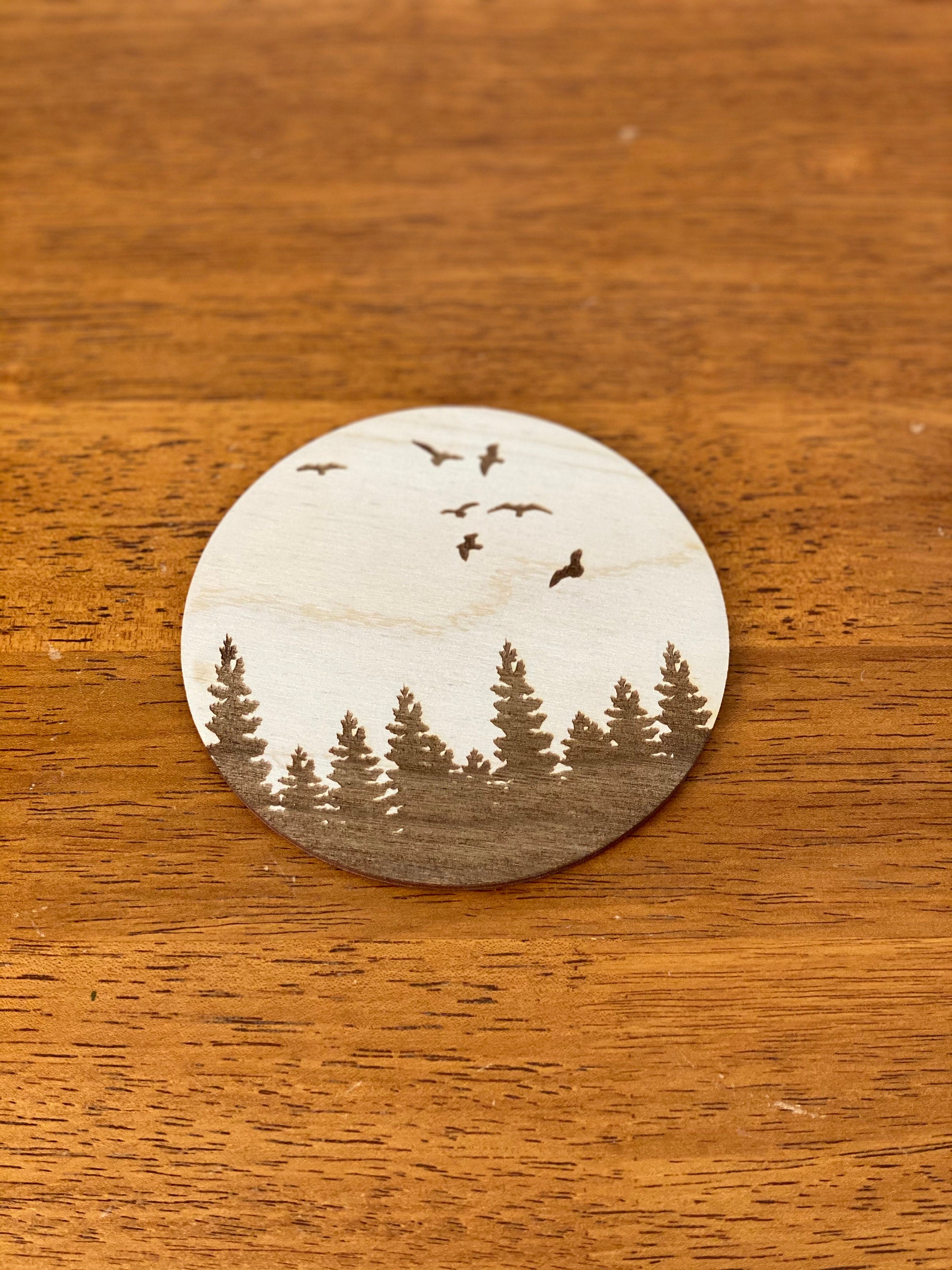 A set of four wooden coasters featuring engraved pine tree and bird designs, showcasing natural wood grain and a rustic aesthetic.