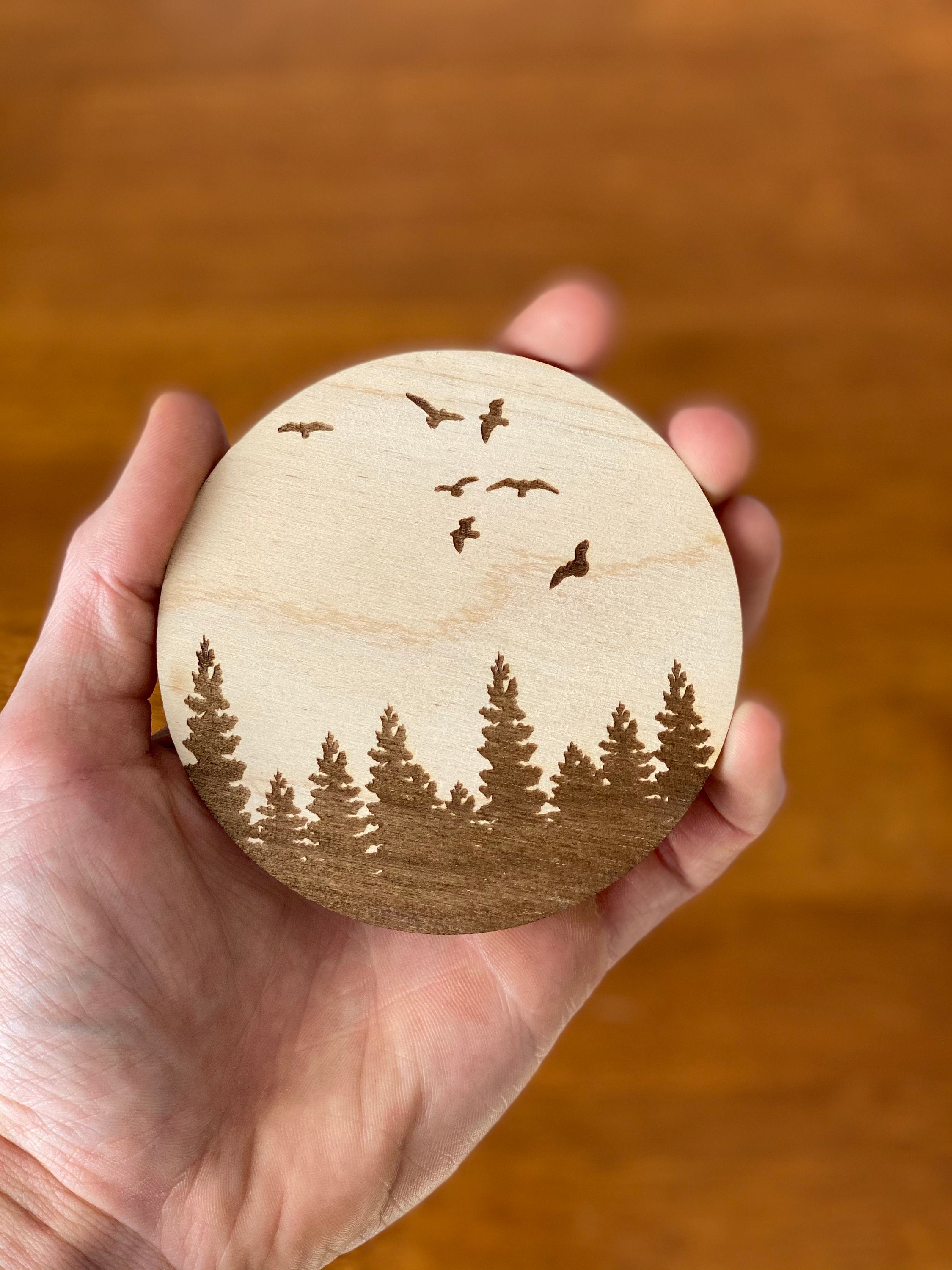 A set of four wooden coasters featuring engraved pine tree and bird designs, showcasing natural wood grain and a rustic aesthetic.