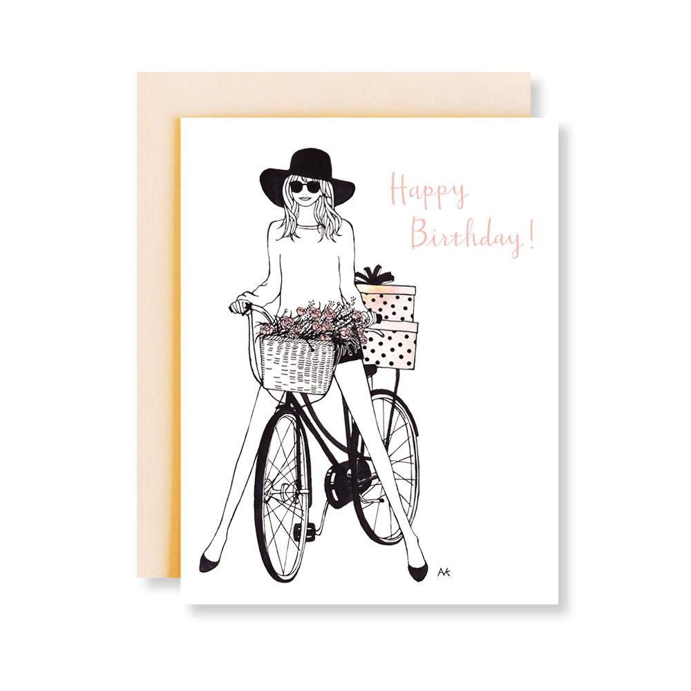A charming birthday card featuring a fashionable girl riding a bicycle, printed on white paper with a peach metallic envelope.