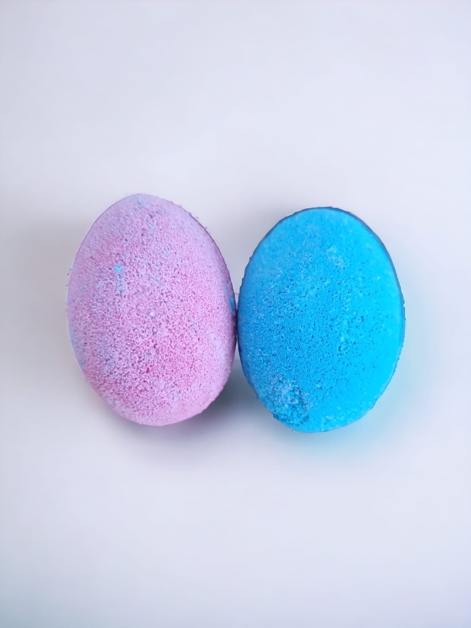 Two colorful egg-shaped bath bombs in purple and blue, packaged in a Kraft bag with an open window, showcasing their delightful design.