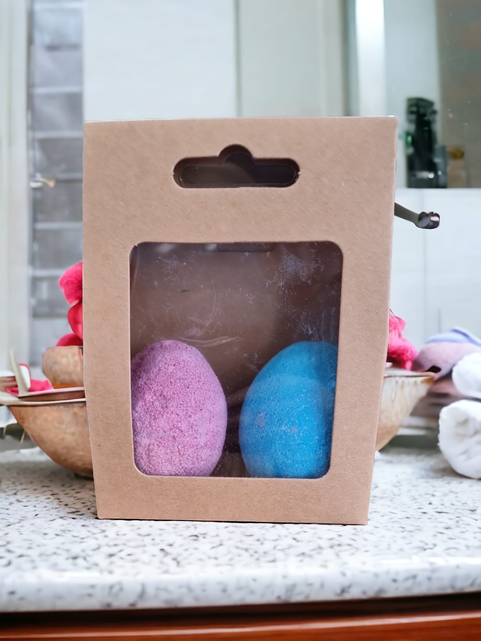 Two colorful egg-shaped bath bombs in purple and blue, packaged in a Kraft bag with an open window, showcasing their delightful design.