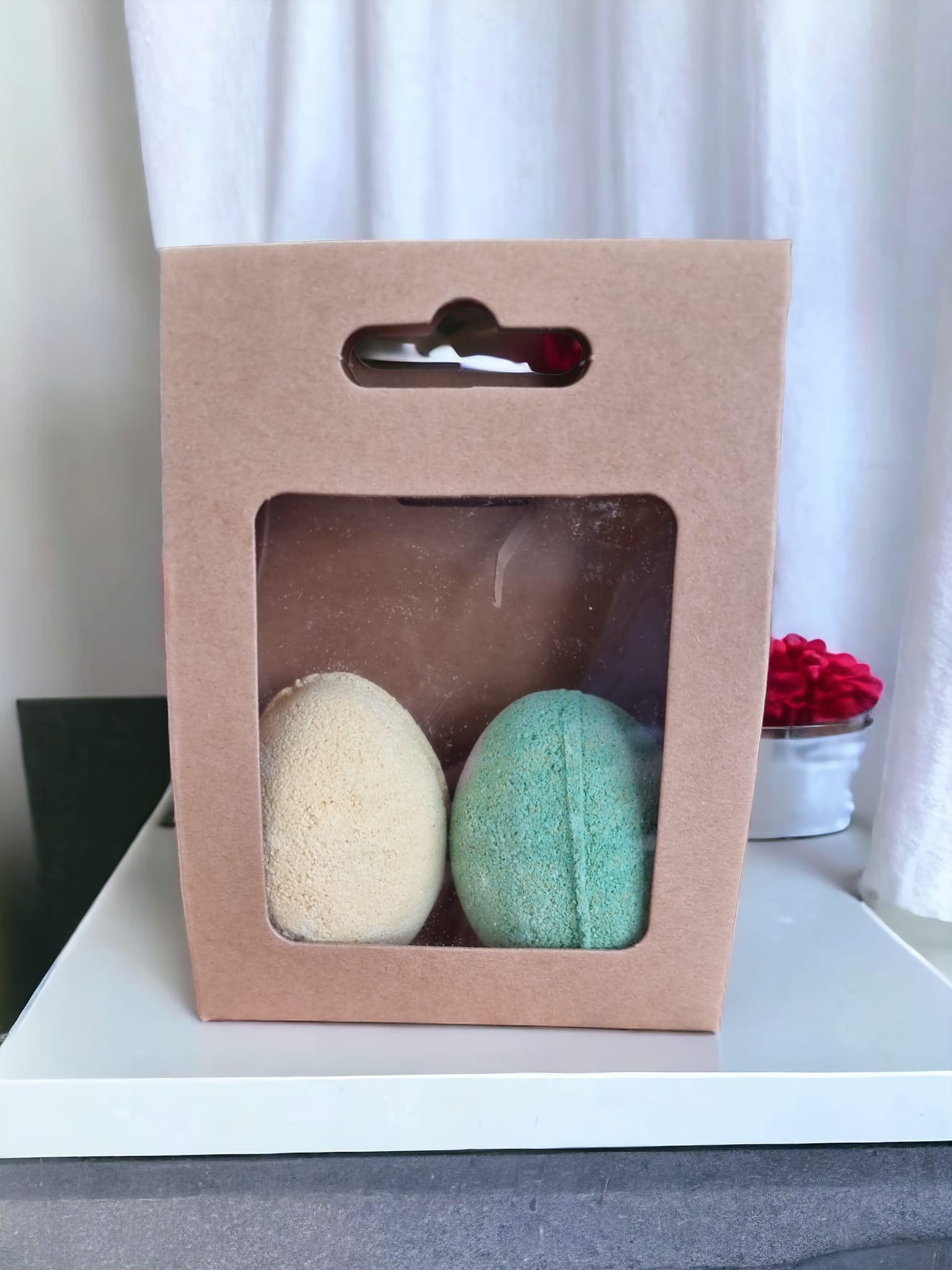 Two vibrant egg-shaped bath bombs in yellow and green, packaged in a Kraft bag with an open window, showcasing their delightful colors.