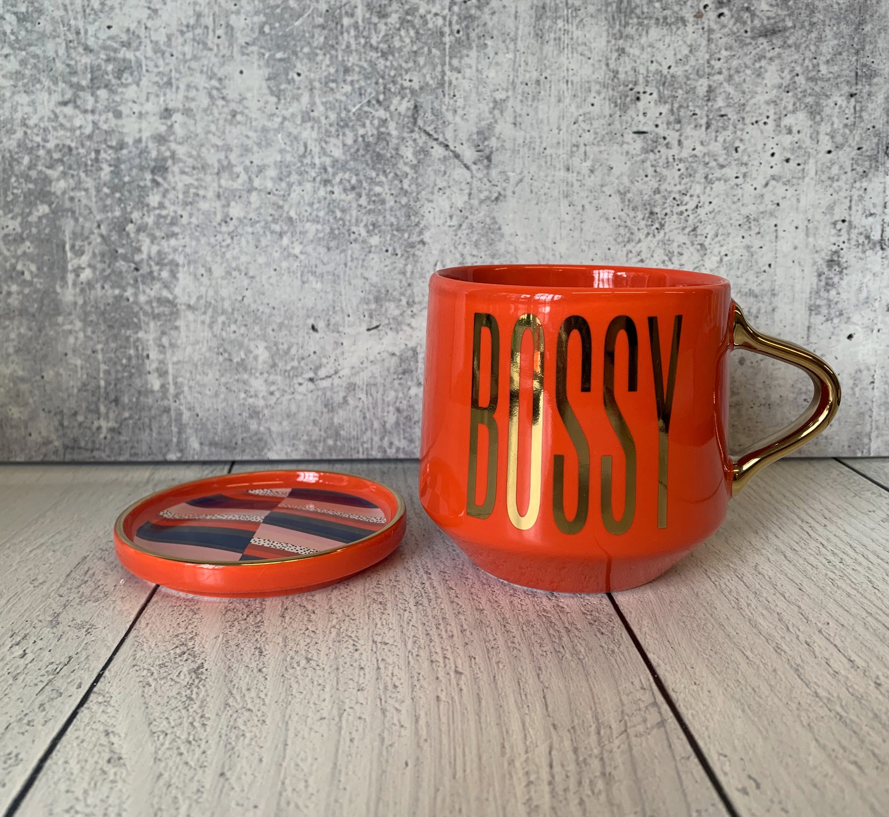 A stylish red and gold ceramic mug with 'BOSSY' printed on it, accompanied by a matching coaster lid.