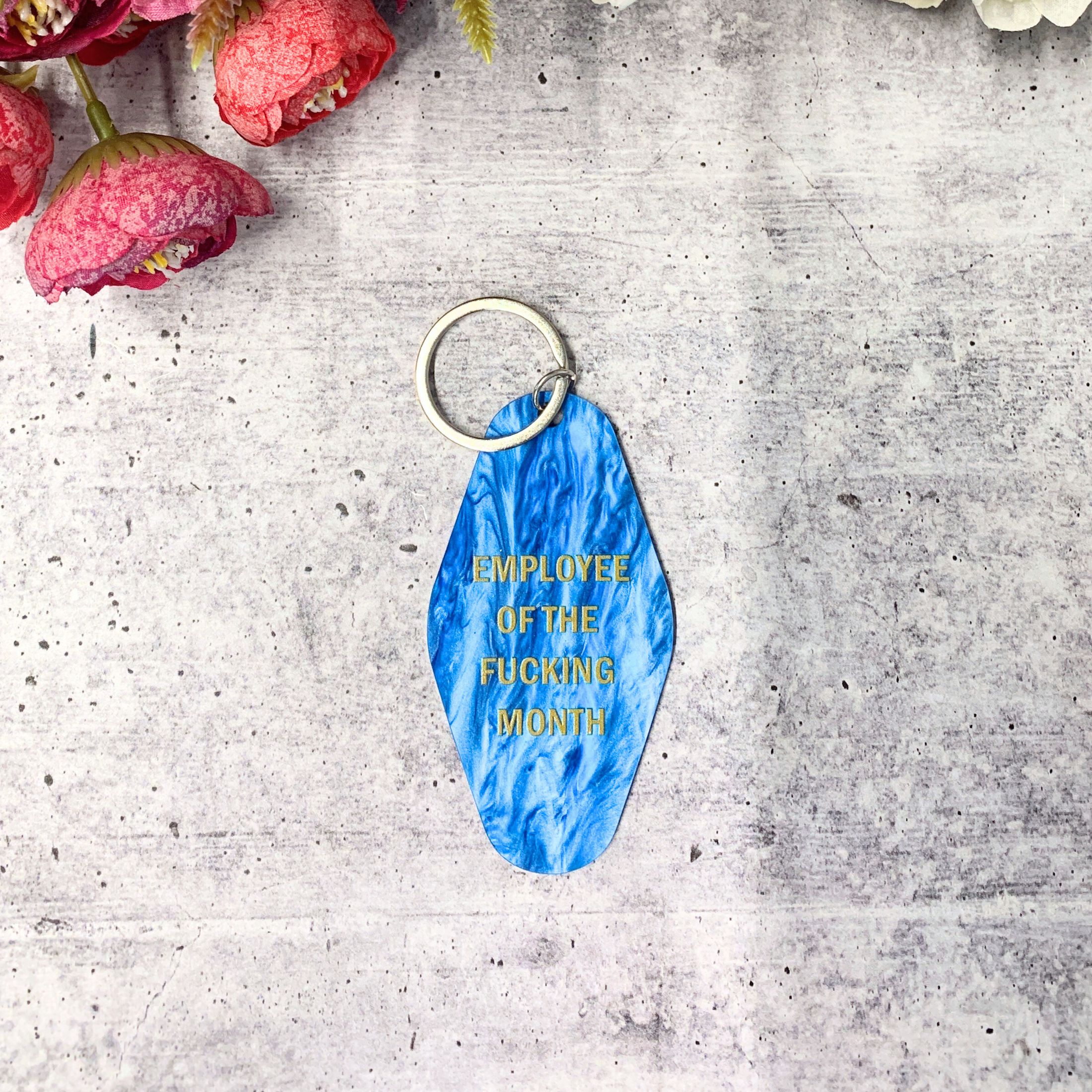 Blue shimmer keychain reading 'EMPLOYEE OF THE FUCKING MONTH' on a split ring, inspired by classic motel key tags.