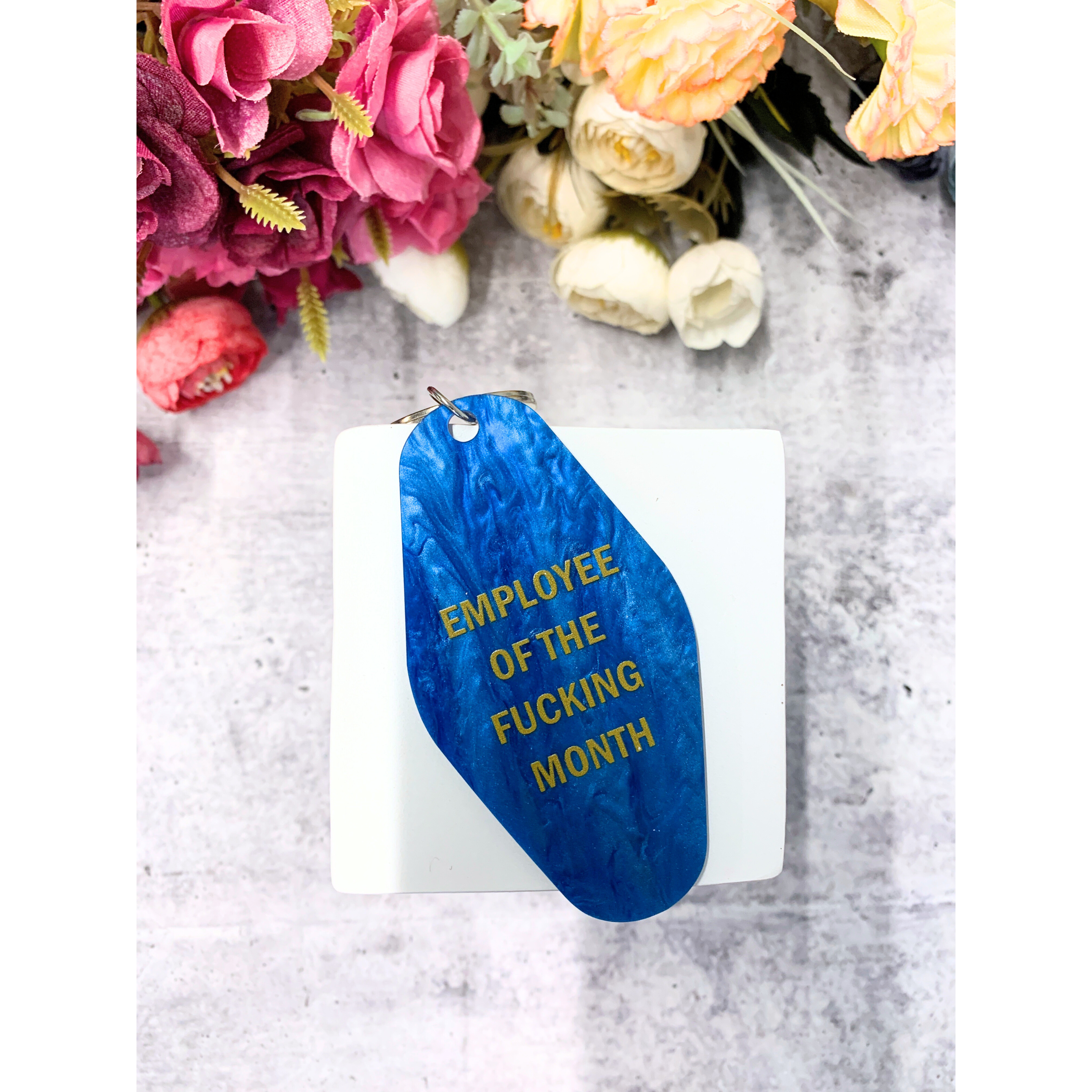 Blue shimmer keychain reading 'EMPLOYEE OF THE FUCKING MONTH' on a split ring, inspired by classic motel key tags.