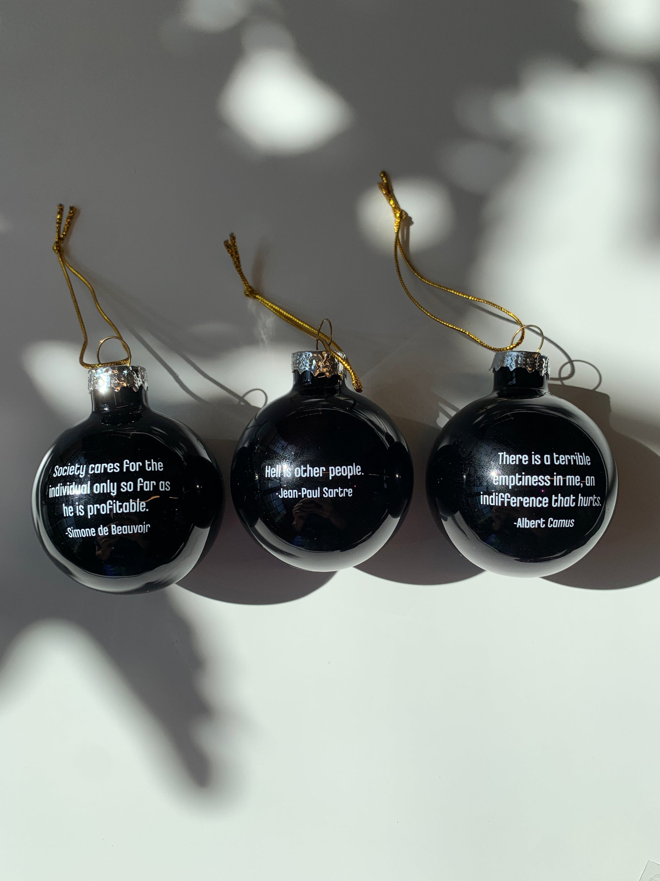 A set of three black glass mini ornaments featuring Albert Camus, Simone de Beauvoir, and Jean-Paul Sartre, perfect for holiday decor.
