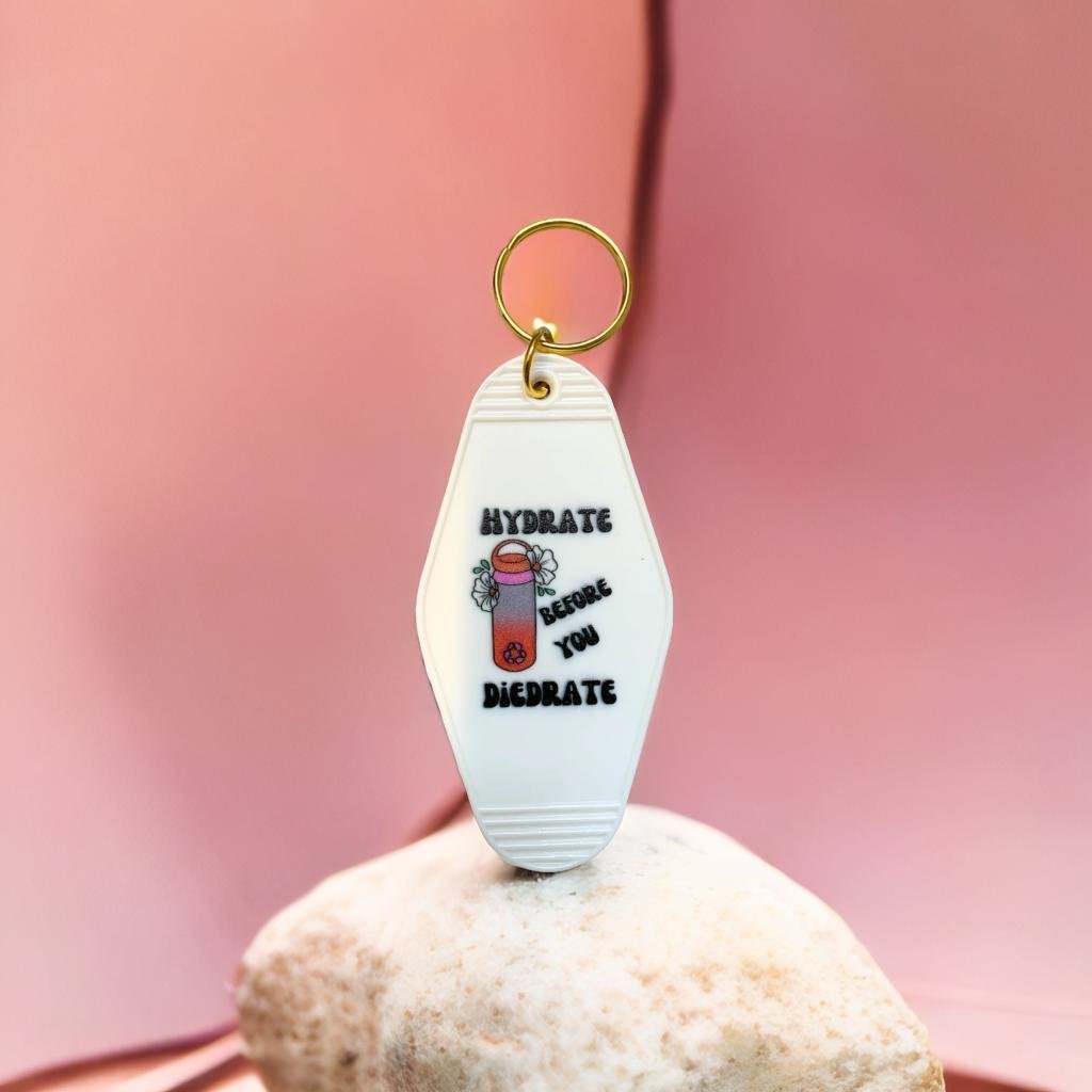 A vibrant motel style keychain with the phrase 'Hydrate Before You Diedrate' in bold letters, featuring a split ring for easy attachment.