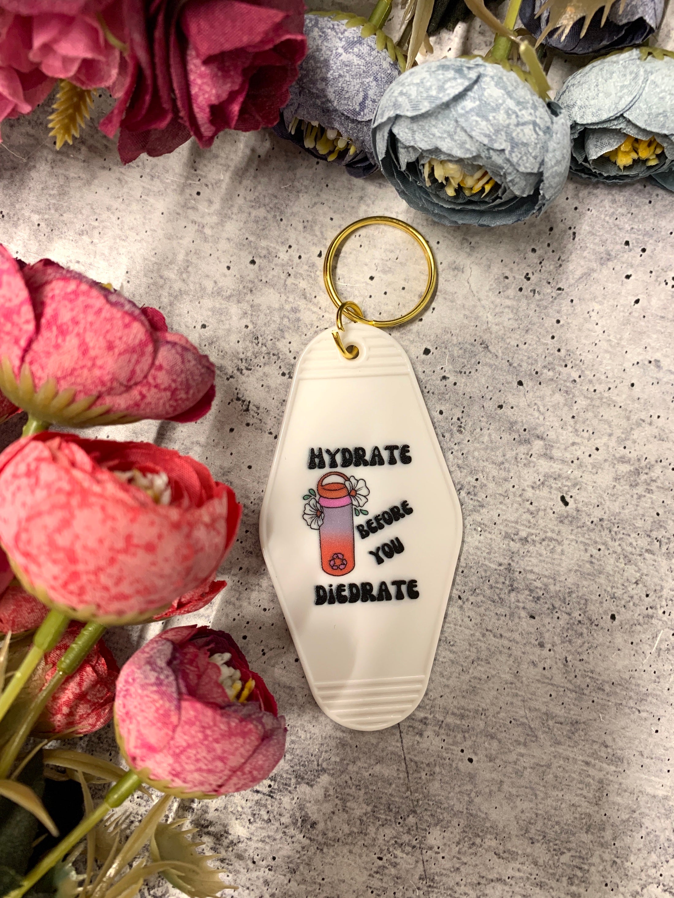 A vibrant motel style keychain with the phrase 'Hydrate Before You Diedrate' in bold letters, featuring a split ring for easy attachment.