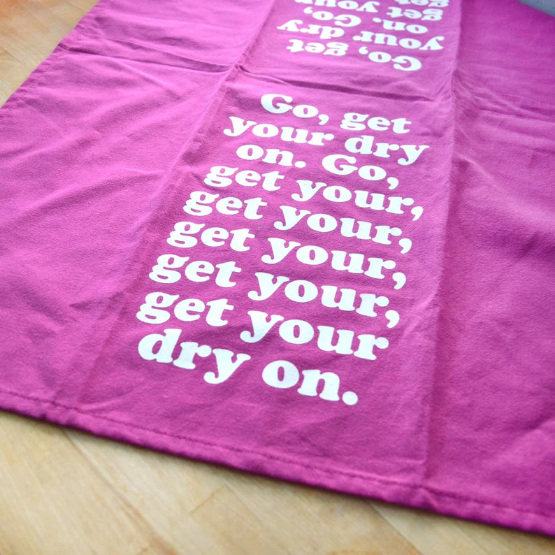 Missy Elliott-inspired cotton tea towel with the phrase 'Go, get your dry on' screen-printed in vibrant colors, hanging on a kitchen rack.
