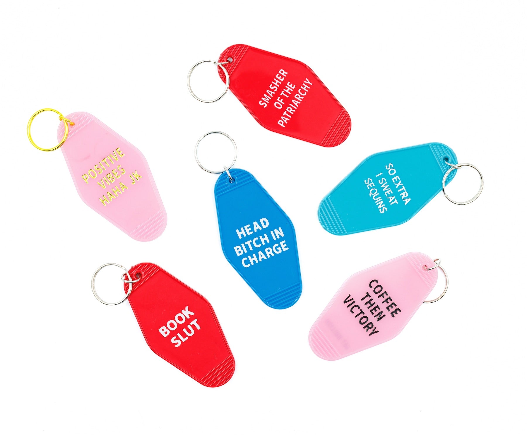 Pink motel keychain with gold lettering reading 'Positive Vibes Haha JK' on the front and 'GET BULLISH' on the back, attached to a brass split ring.