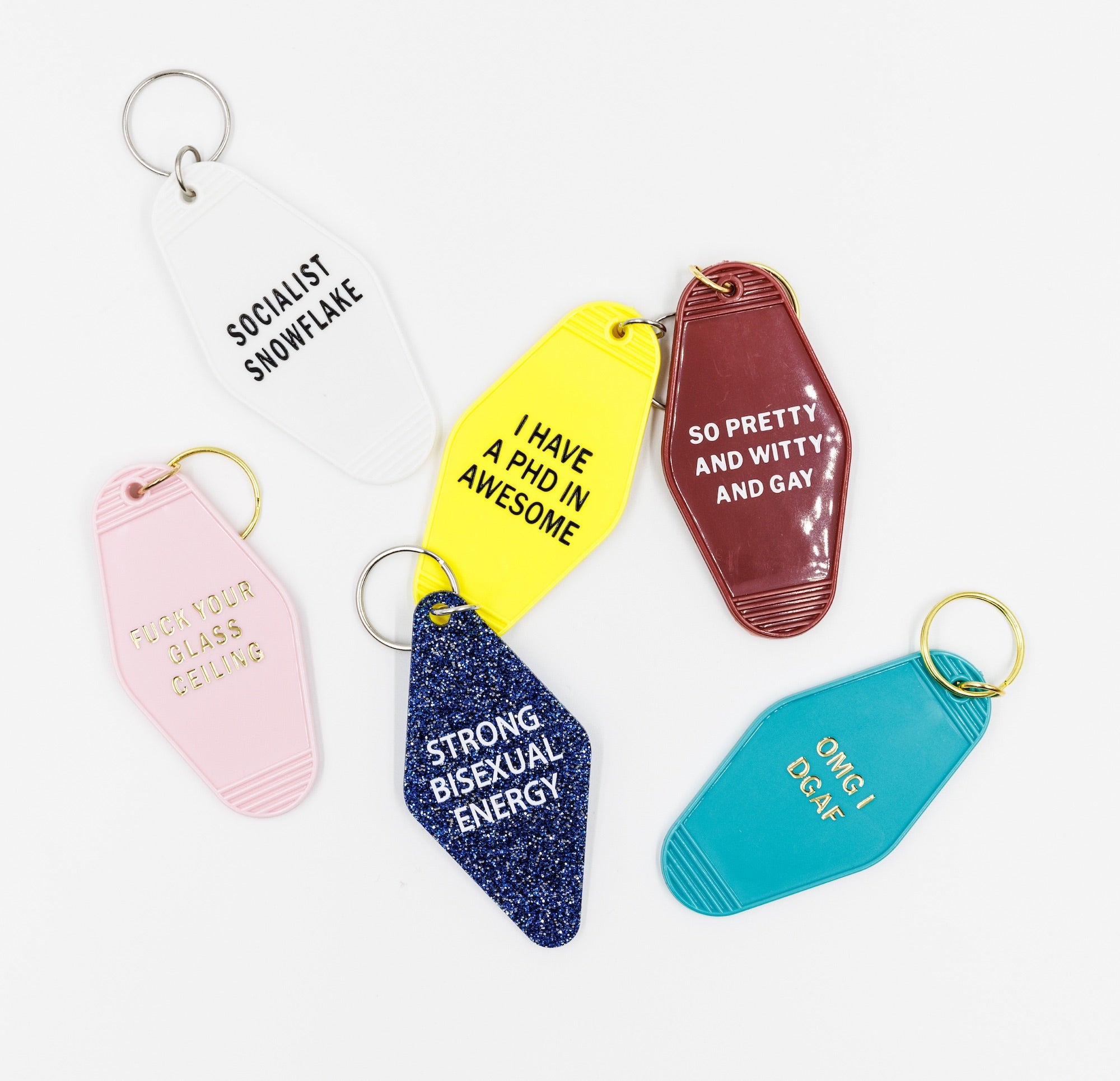 A vibrant dark red keychain with white lettering that reads 'SO PRETTY AND WITTY AND GAY', featuring a classic motel key tag design.