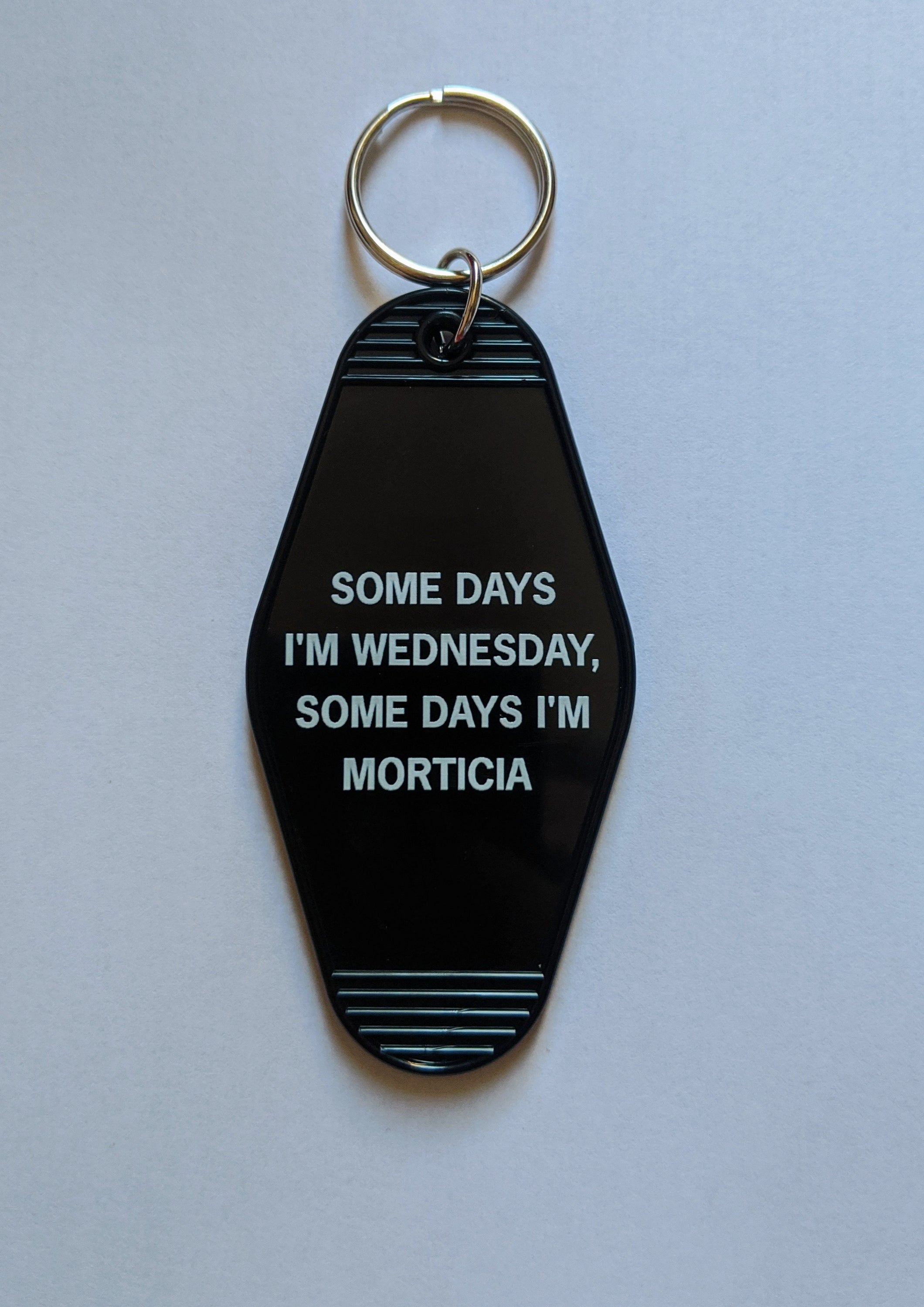 Black motel-style keychain with white lettering reading 'Some Days I'm Wednesday, Some Days I'm Morticia', featuring a split ring.