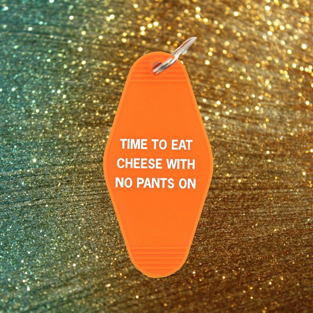 Orange motel-style keychain with white text reading 'TIME TO EAT CHEESE WITH NO PANTS ON', featuring a split ring attachment.