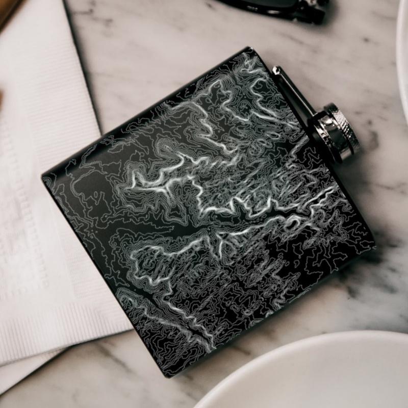 Matte black hip flask featuring a custom engraved map of Zion National Park, Utah, with coordinates, alongside a canvas bag and funnel.