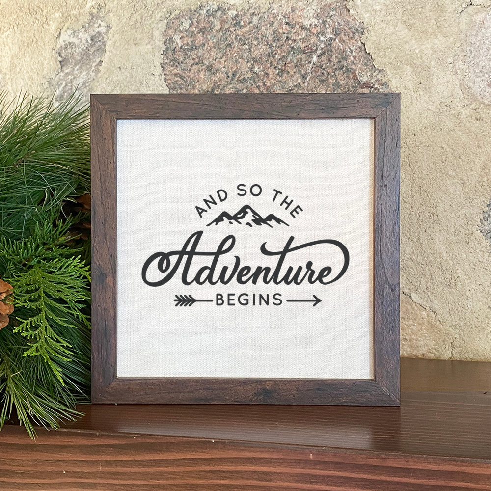 Framed sign reading 'And so the Adventure Begins' with a stylish wood frame and linen-look background.