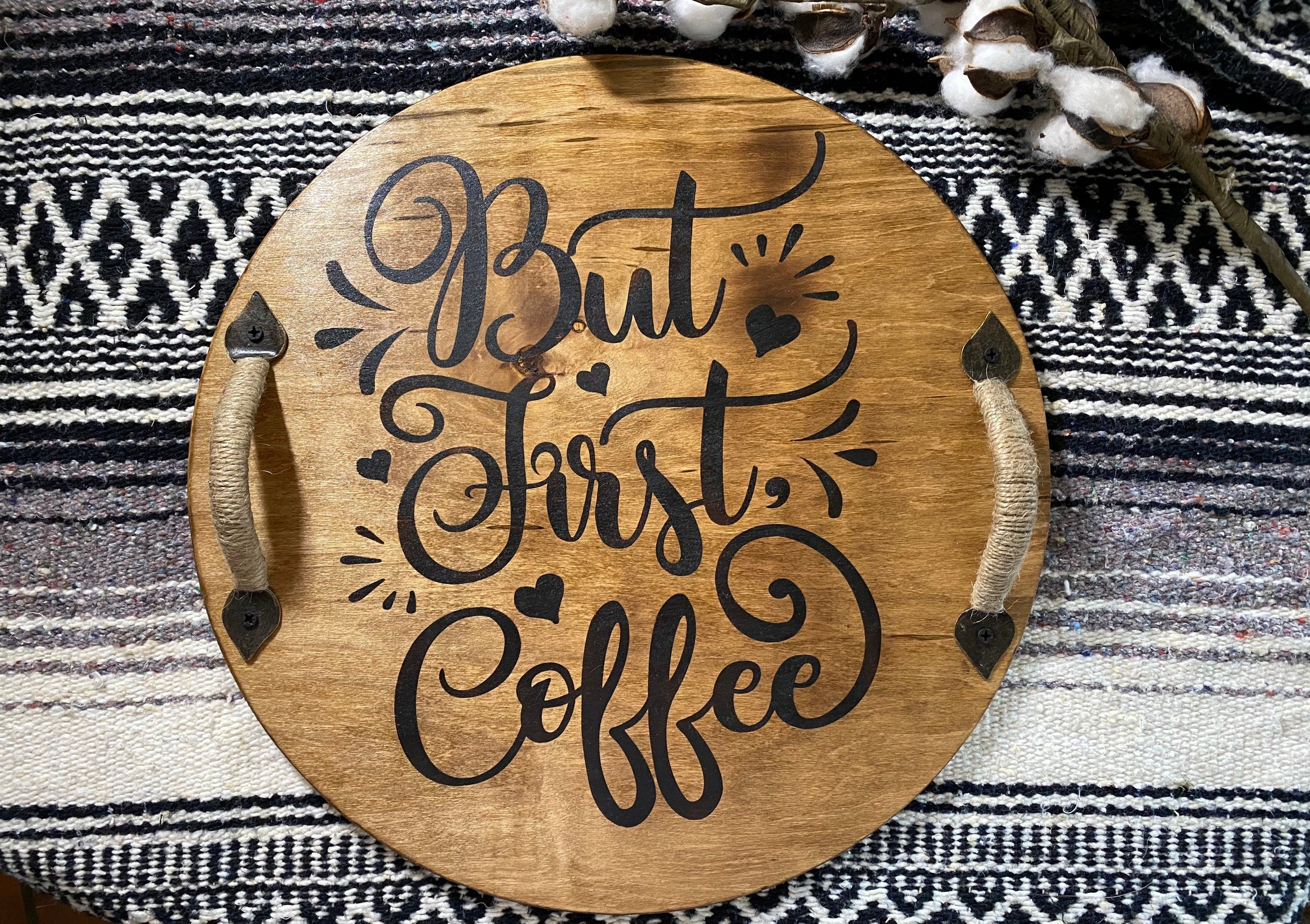 A rustic wooden serving tray with 'But First Coffee' burned into the surface, featuring black metal handles and a rich chestnut stain.