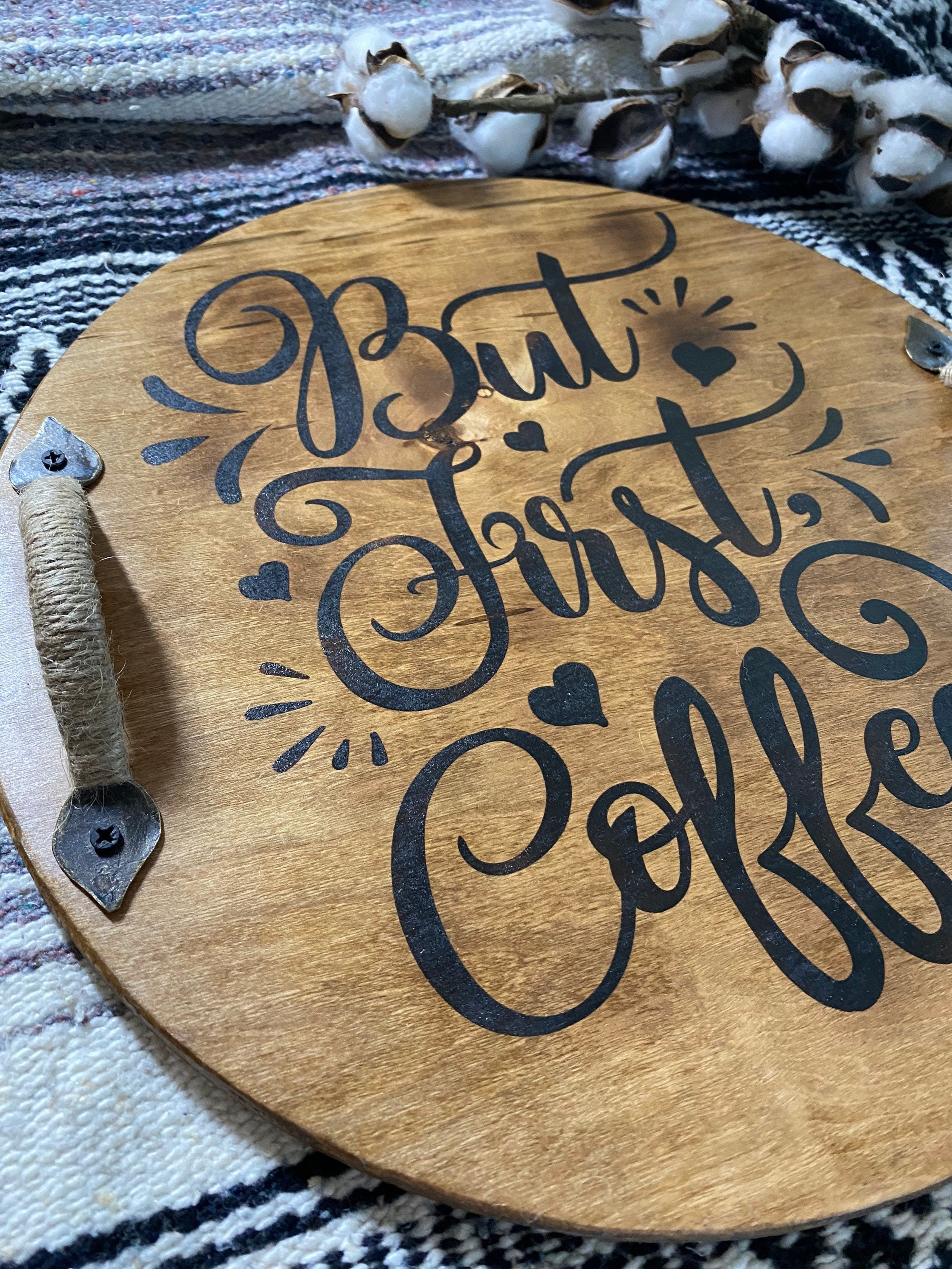 A rustic wooden serving tray with 'But First Coffee' burned into the surface, featuring black metal handles and a rich chestnut stain.