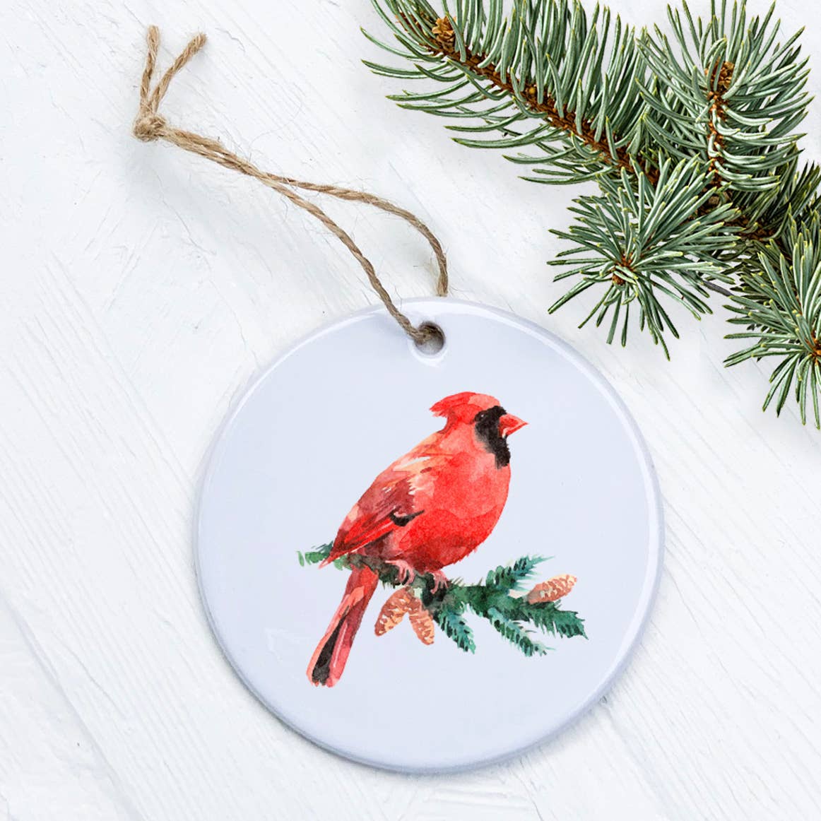 A beautiful Cardinal perched on an evergreen branch, featured on a high-quality porcelain ornament with a glossy finish.