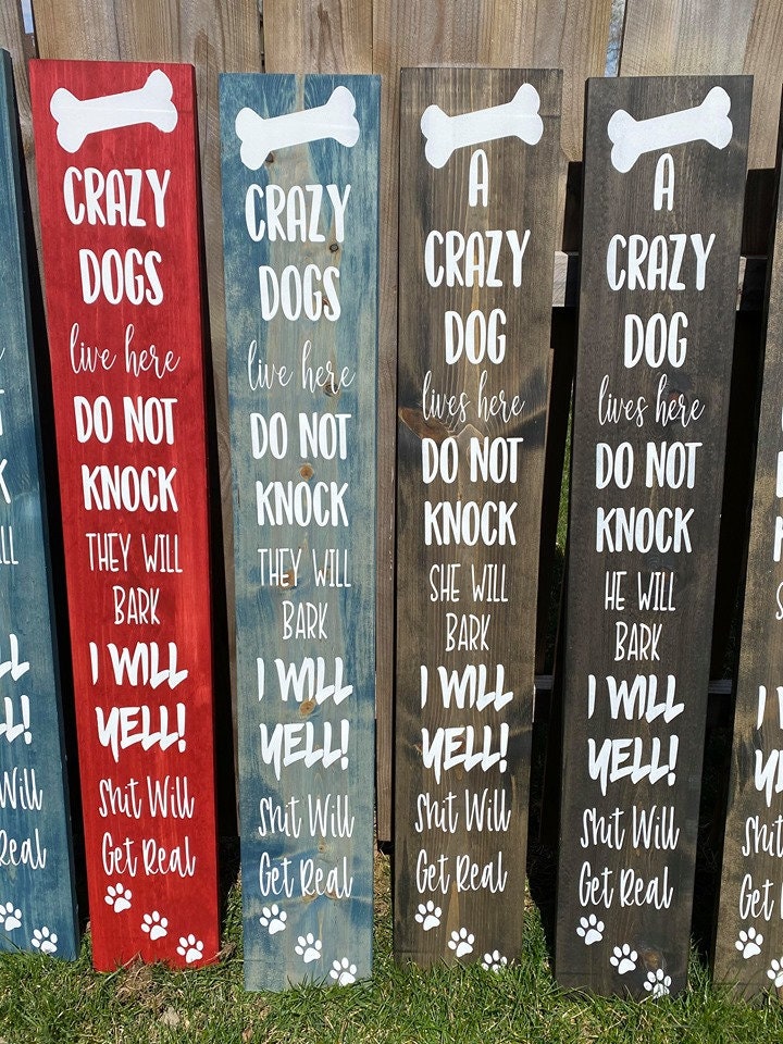 A humorous wooden front door sign that reads 'Crazy Dogs Live Here Do Not Knock They Will Bark I Will Yell Shit Will Get Real', showcasing unique wood grain.