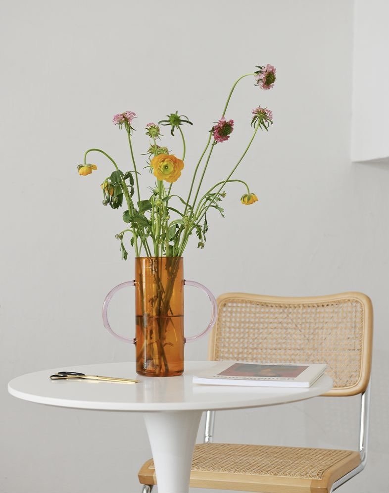 A whimsical dual-handle glass flower vase, handblown with a unique design, perfect for seasonal flowers and elegant table settings.