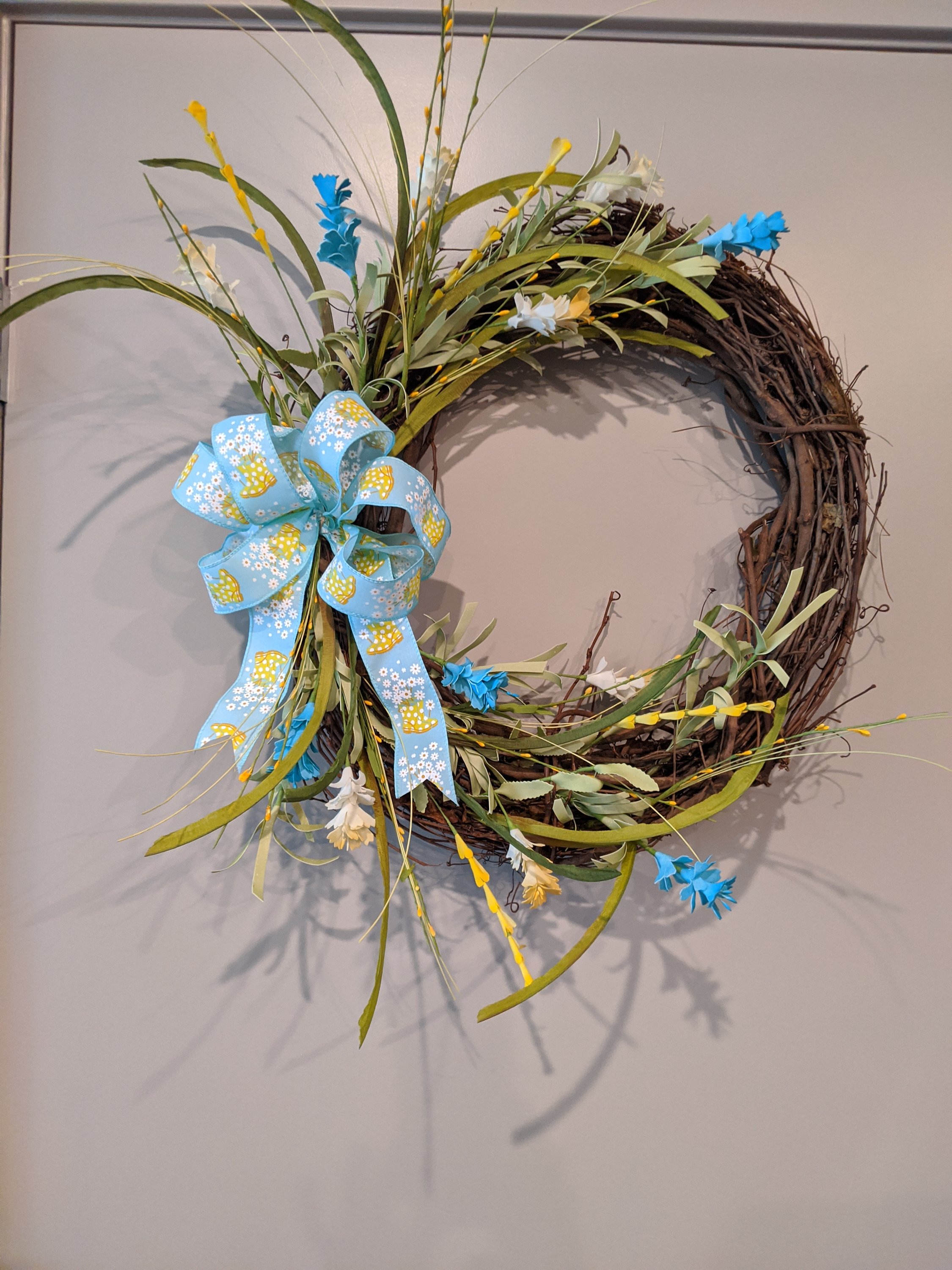 A vibrant 18-inch handmade grapevine wreath featuring aquas and yellows, adorned with a unique rainboot and floral ribbon, perfect for spring decor.