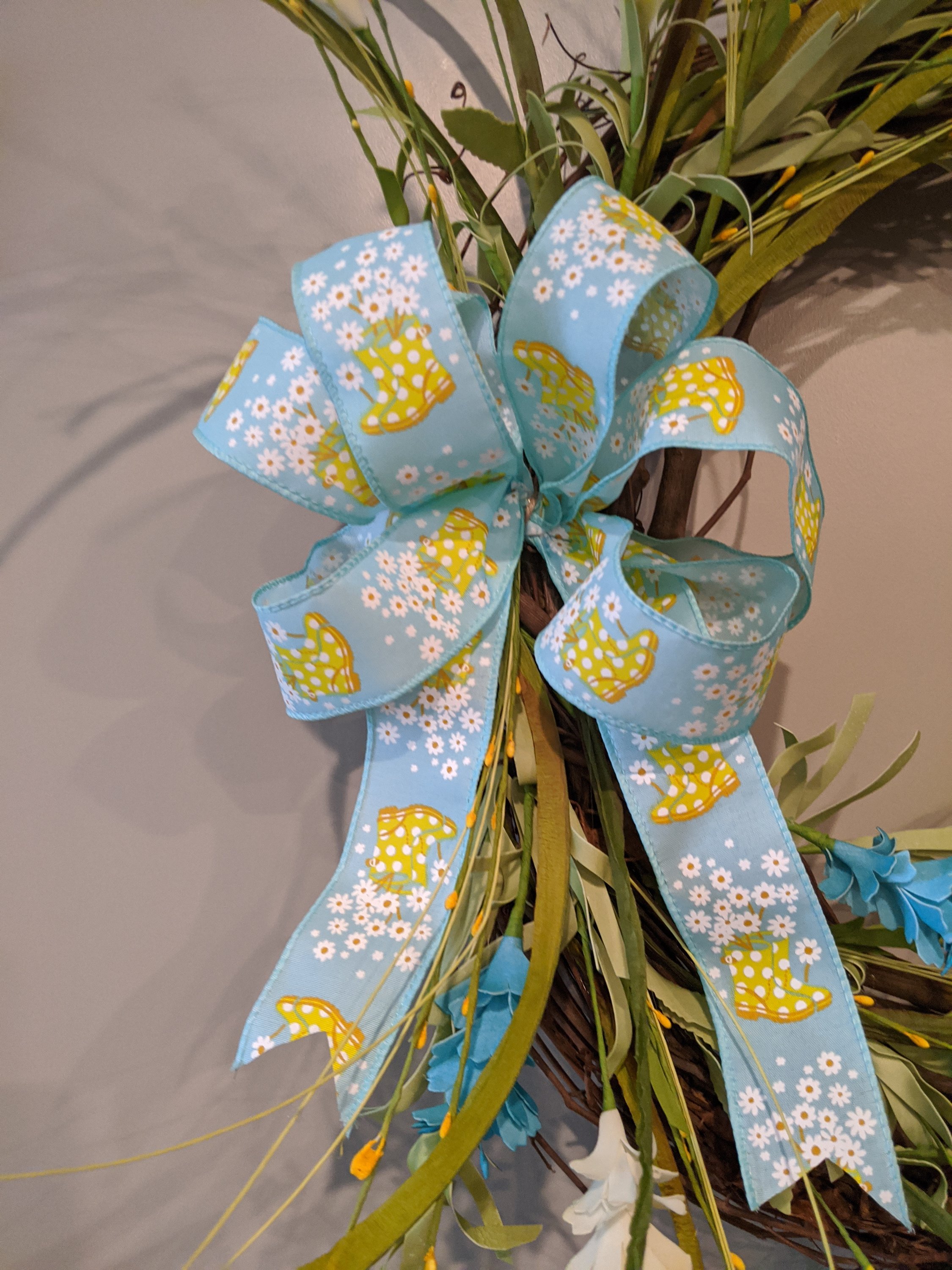 A vibrant 18-inch handmade grapevine wreath featuring aquas and yellows, adorned with a unique rainboot and floral ribbon, perfect for spring decor.