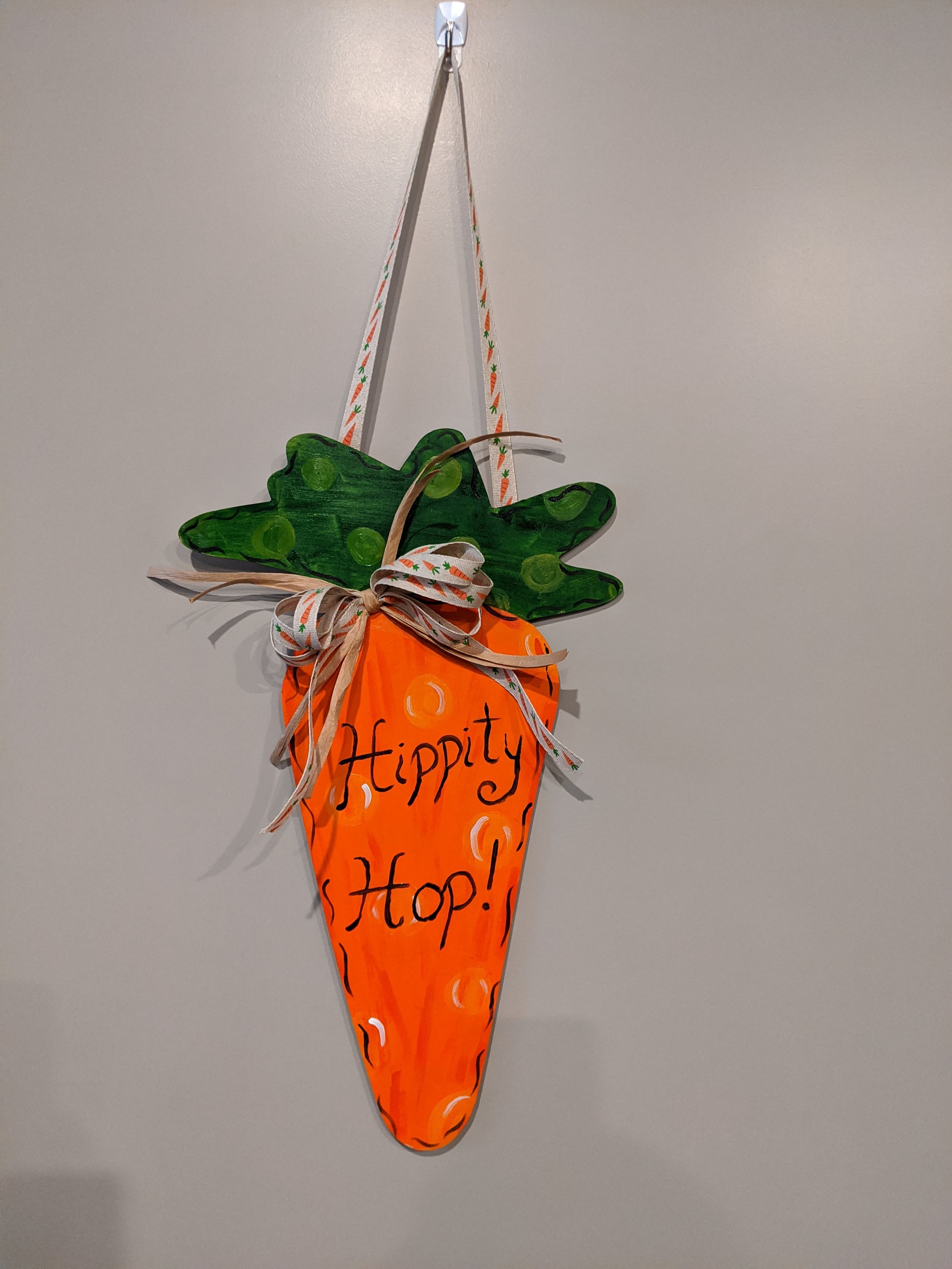 Handpainted wooden hippity hop carrot door hanger with vibrant colors and a charming carrot ribbon accent, perfect for spring and Easter decor.