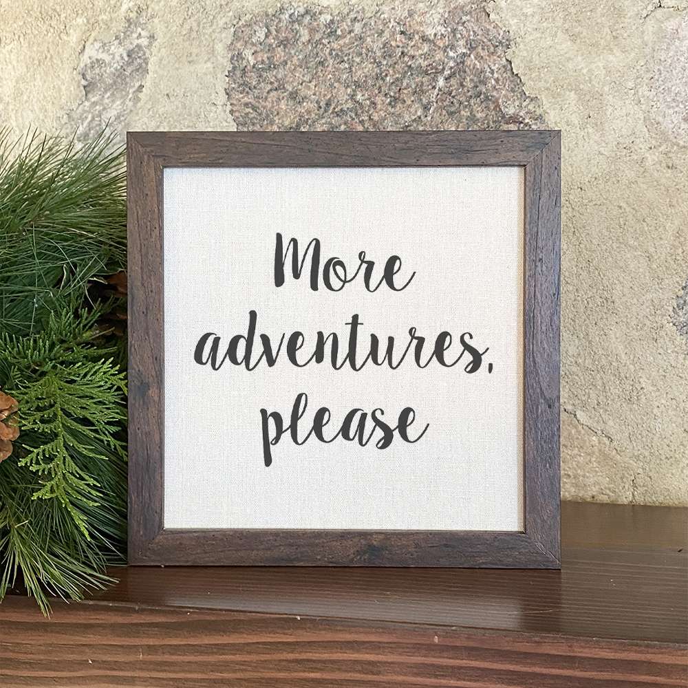 A beautifully framed sign that reads 'More Adventures Please', featuring a natural wood frame and a linen-look neutral background.