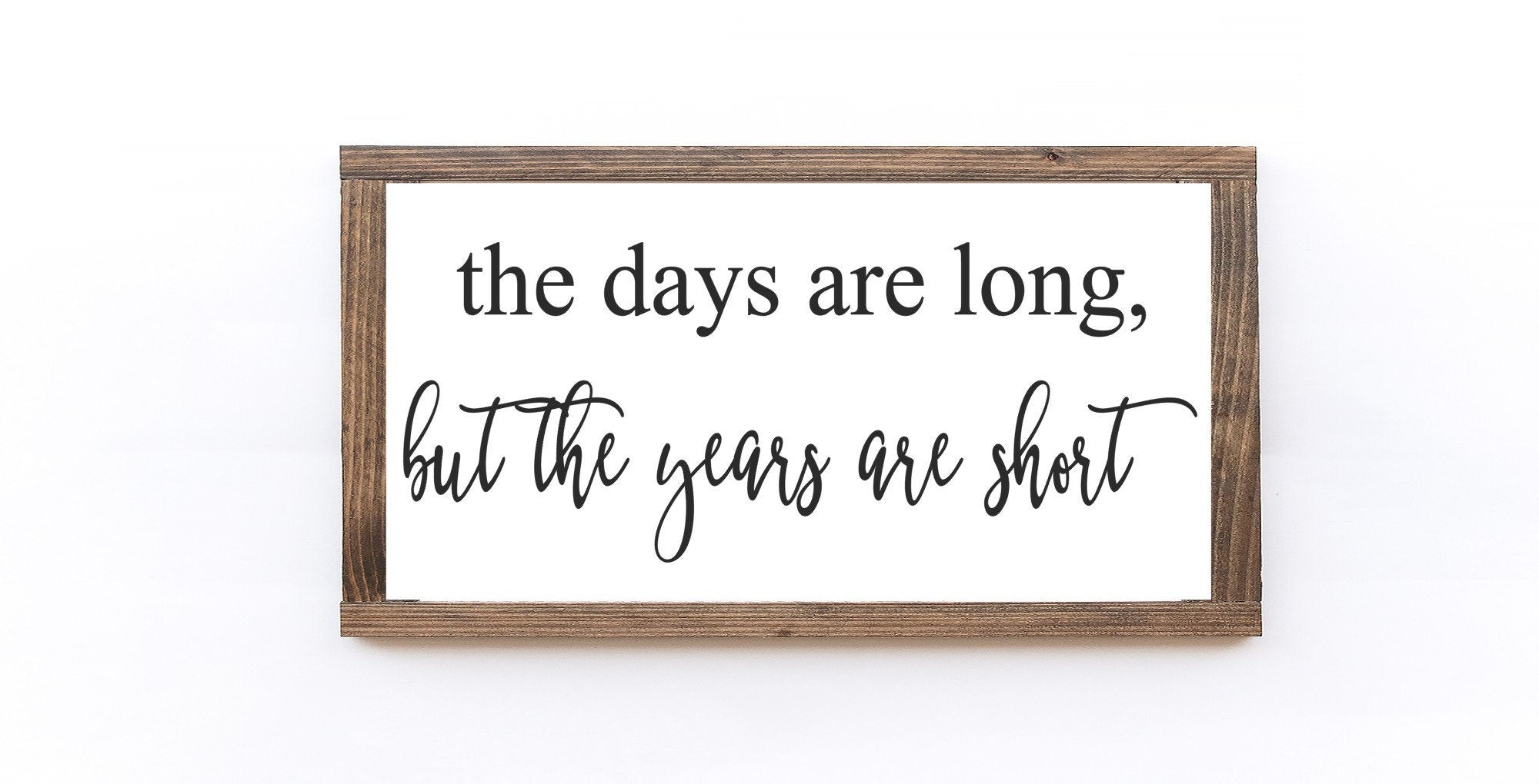 A handmade wooden sign featuring the phrase 'The Days Are Long, But The Years Are Short' in elegant lettering on a matte white background.